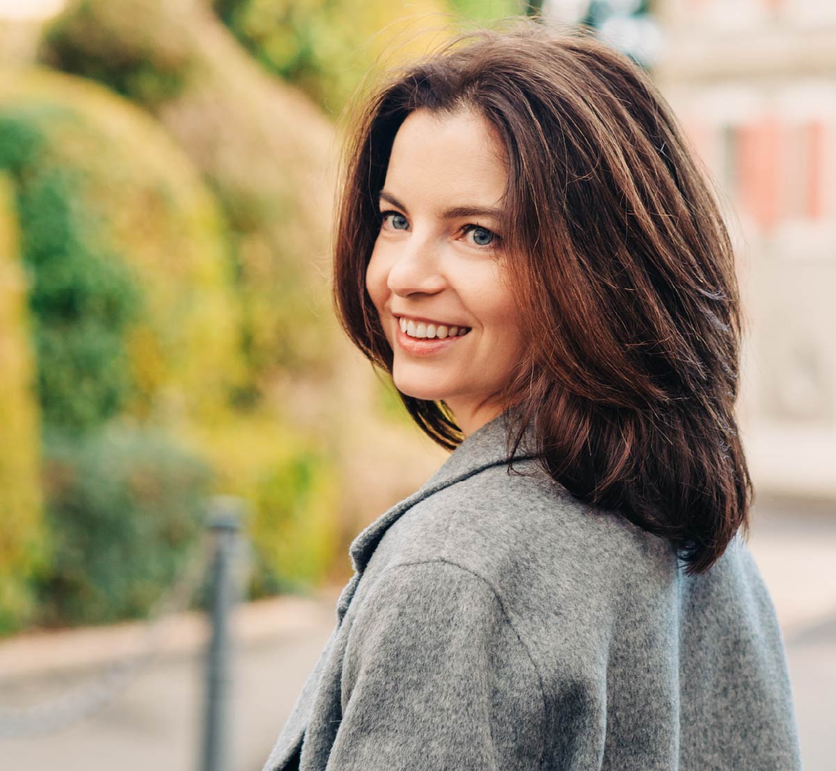 A woman looking over her shoulder while standing in front of a beautiful house and topiary.