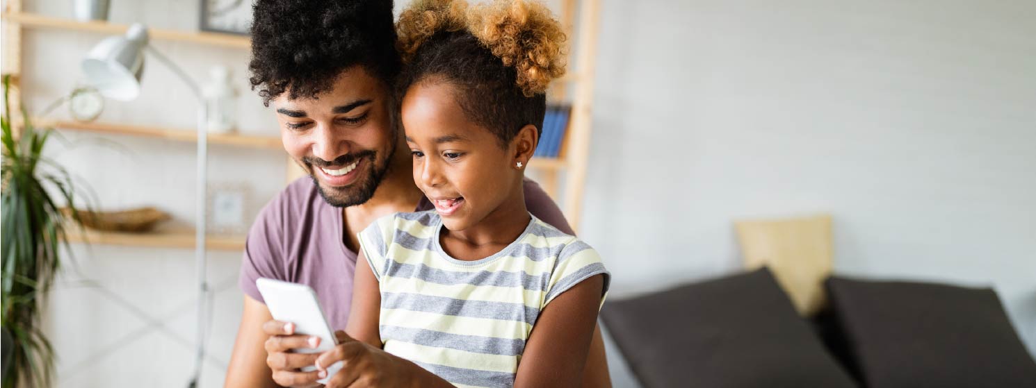 A father and daughter sitting together, smiling, and looking at a phone screen.