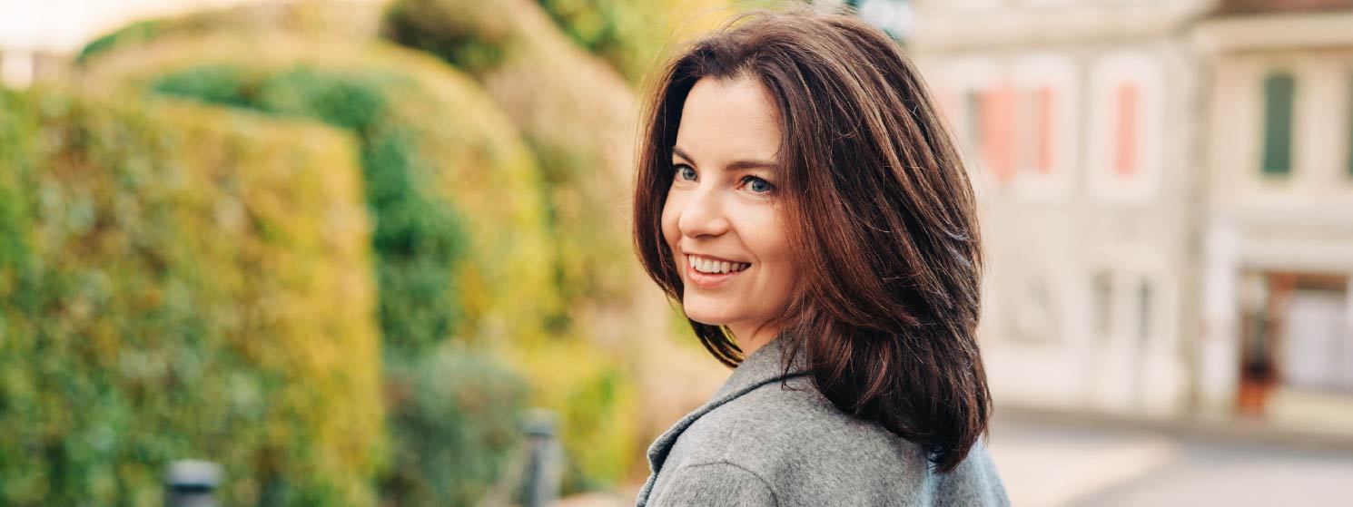 A woman looking over her shoulder while standing in front of a beautiful house and topiary.