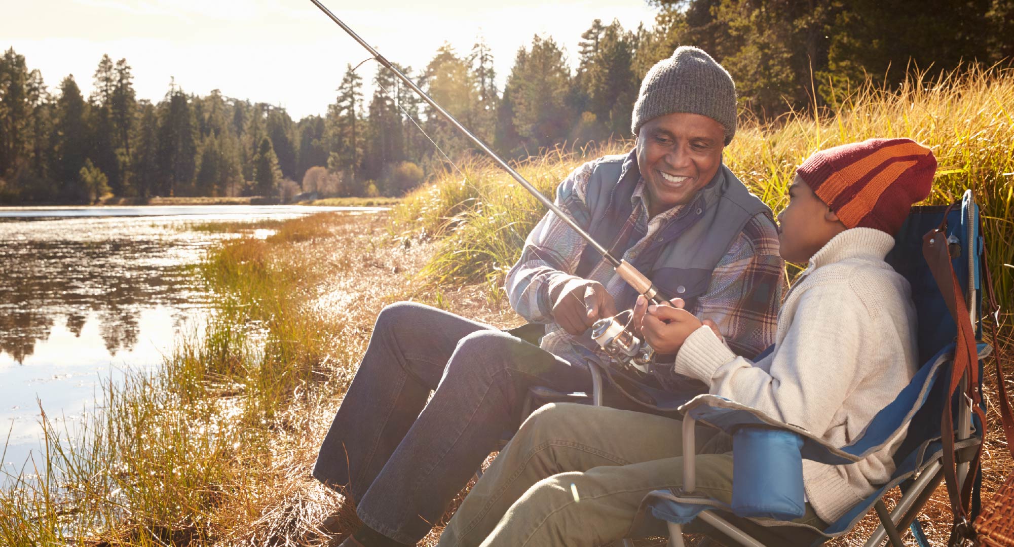 A grandfather and grandson sitting on a lake shore and fishing.